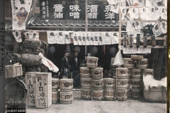 Epiciers à l'entrée de leur boutique, devant laquelle sont exposés des fûts de bière, de «miso» (pâte de haricots de soja fermentée), de saké et du charbon de bois, Tôkyô, Japon, 1926-1927, (Autochrome, 9 x 12 cm), Roger Dumas, Département des Hauts-de-Seine, musée Albert-Kahn, Archives de la Planète, A 68 627 X